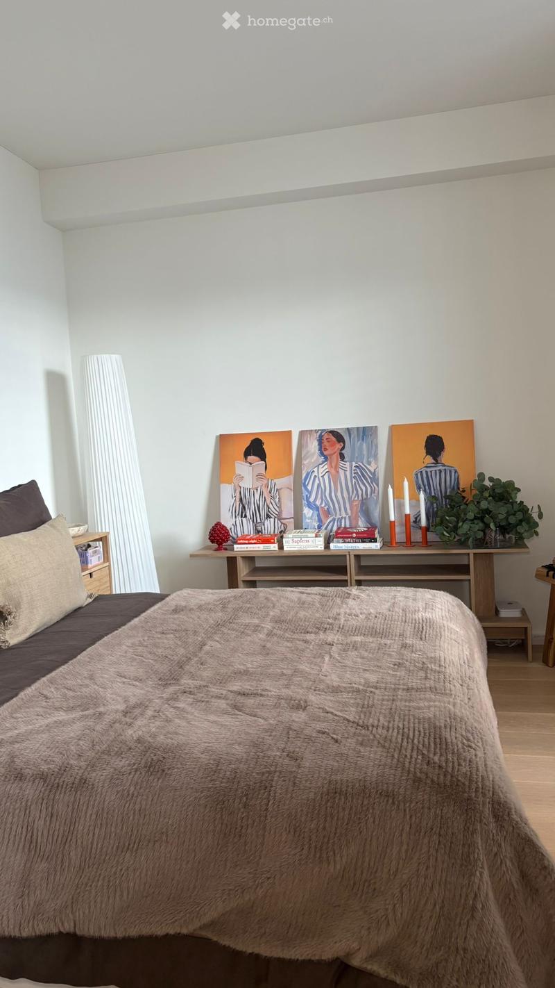 A modern bedroom with a large bed covered in a soft, light brown fur throw, a wooden nightstand with a lamp, a small table with books, candles, and artwork, and a potted plant on the floor.