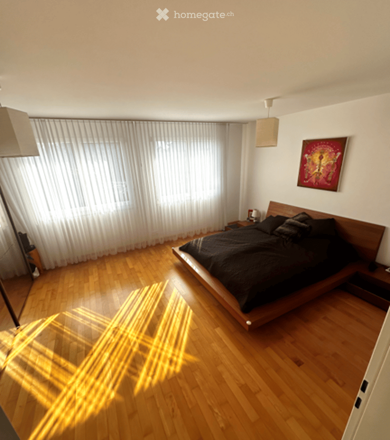 Modern bedroom with wooden floor, large window with white curtains, platform bed with dark bedding, nightstand, lamp, and artwork on wall.