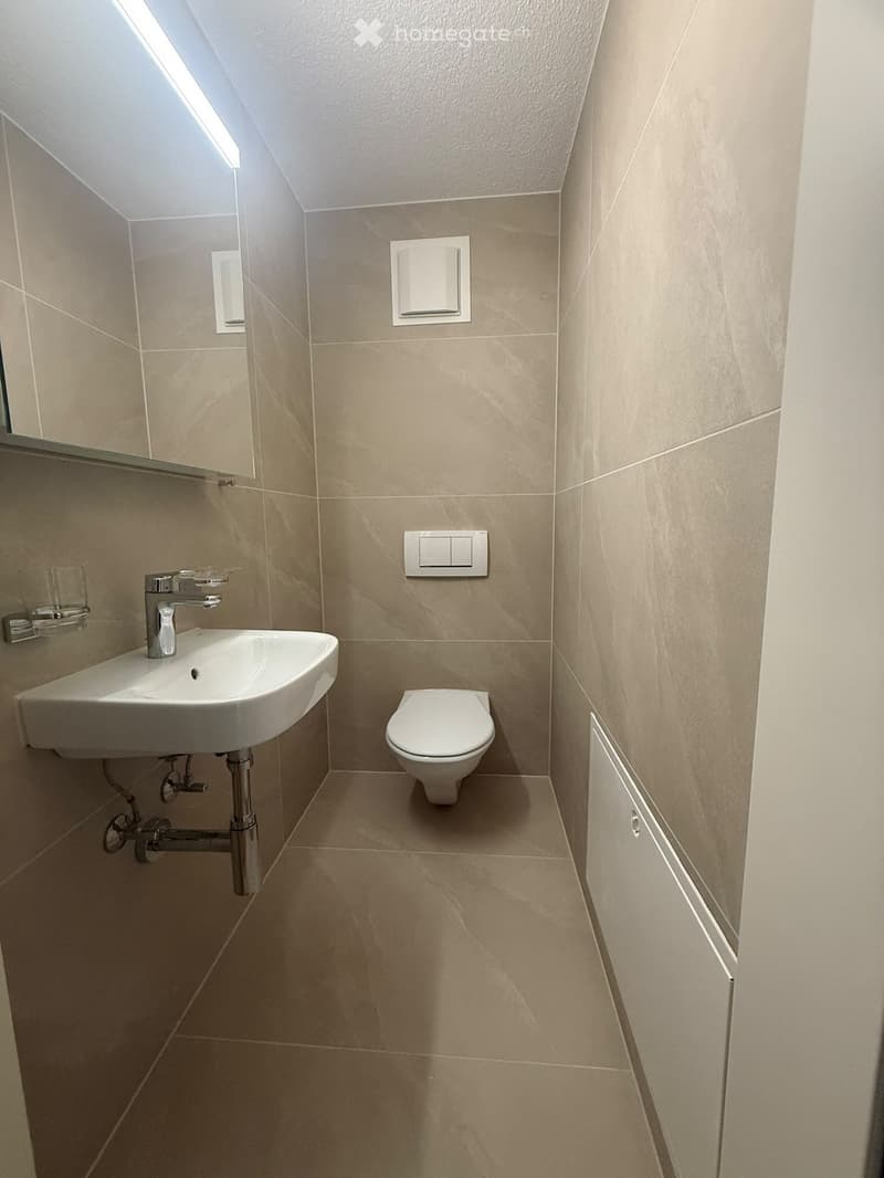 Modern bathroom with beige tiles, a white sink with a chrome faucet, and a white toilet. The room features a mirrored cabinet and recessed lighting.