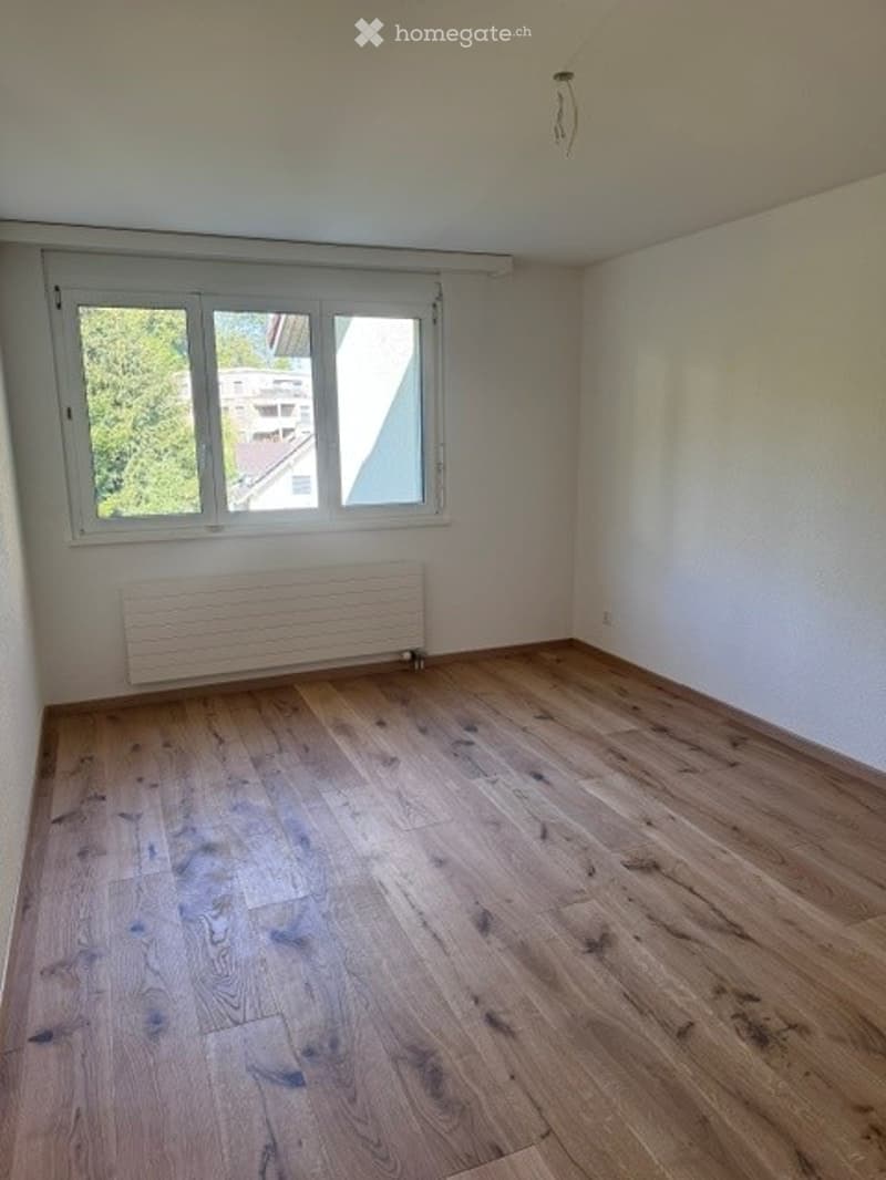 Empty room with wooden flooring, white walls, and a single window with a radiator below it.