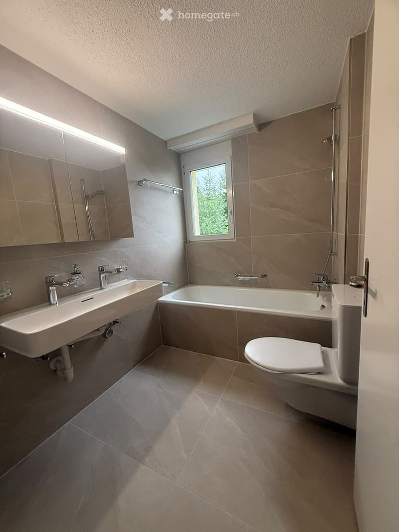 Modern bathroom with a double sink, bathtub, toilet, and large mirror. Features tiled walls and floor, and a window with a view of greenery.