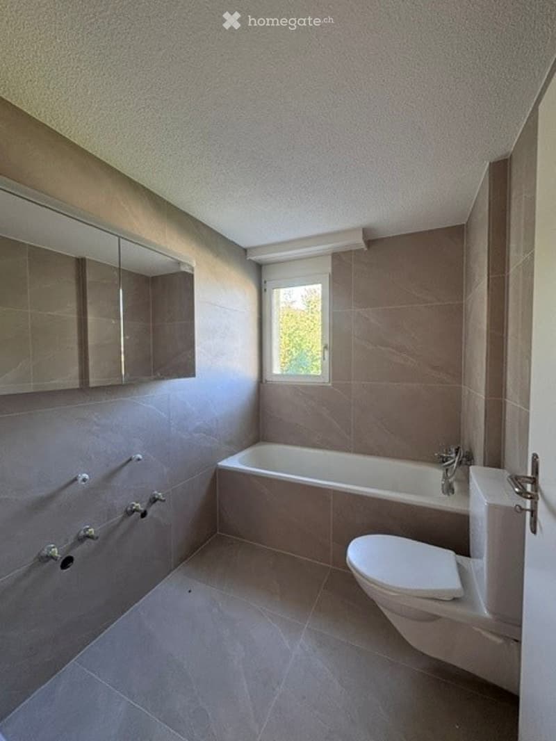 Modern bathroom with a bathtub, toilet, and a large mirror above a sink. The walls and floor are tiled in a neutral color scheme, and there is a window providing natural light.