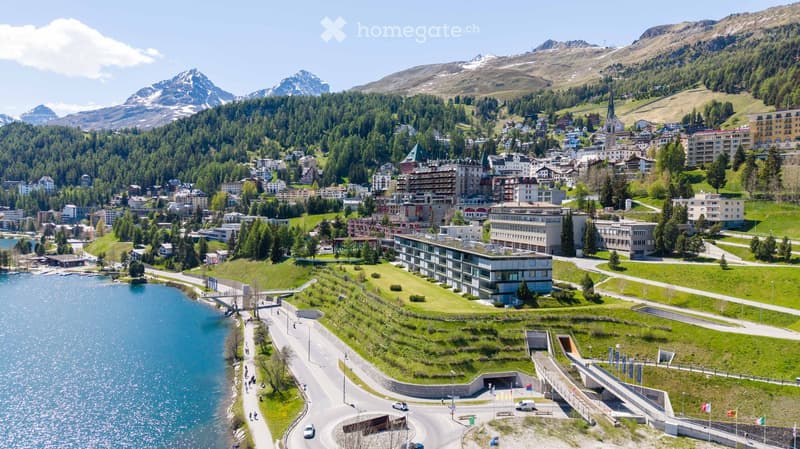 A scenic view of a town nestled in a valley surrounded by mountains, with a lake in the foreground and various buildings, including hotels and residential structures, scattered throughout the landscape.