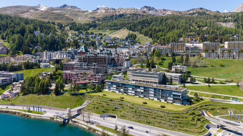 Aerial view of a town nestled in a valley surrounded by mountains, featuring a mix of residential and commercial buildings, green spaces, and a lake in the foreground.