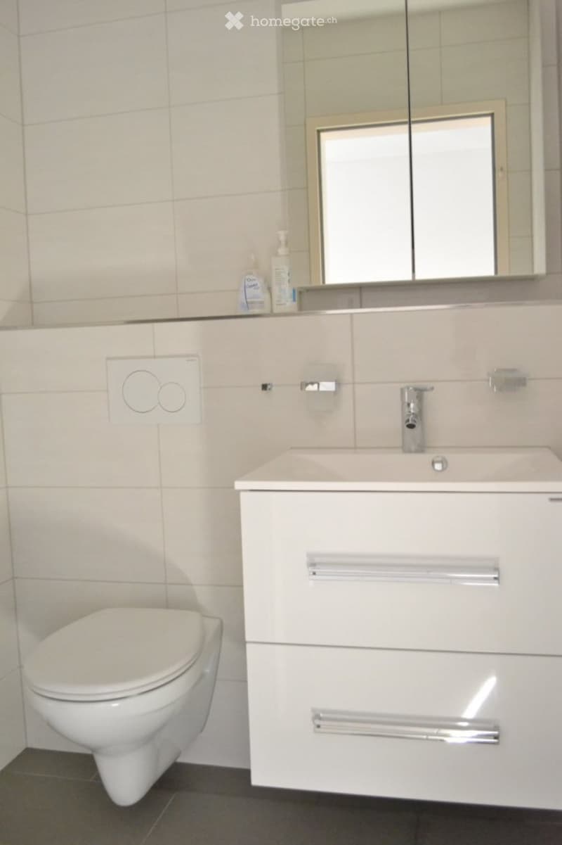 Modern bathroom with white tiles, a toilet, a sink with a modern faucet, a mirrored cabinet, and a sleek white cabinet with silver handles.