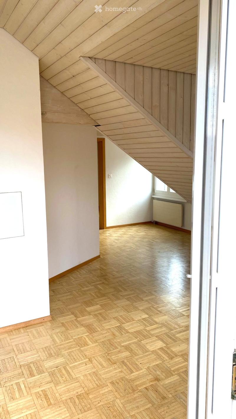 Empty room with wooden herringbone flooring, white walls, and exposed wooden ceiling beams. A small window and radiator are visible in the background.