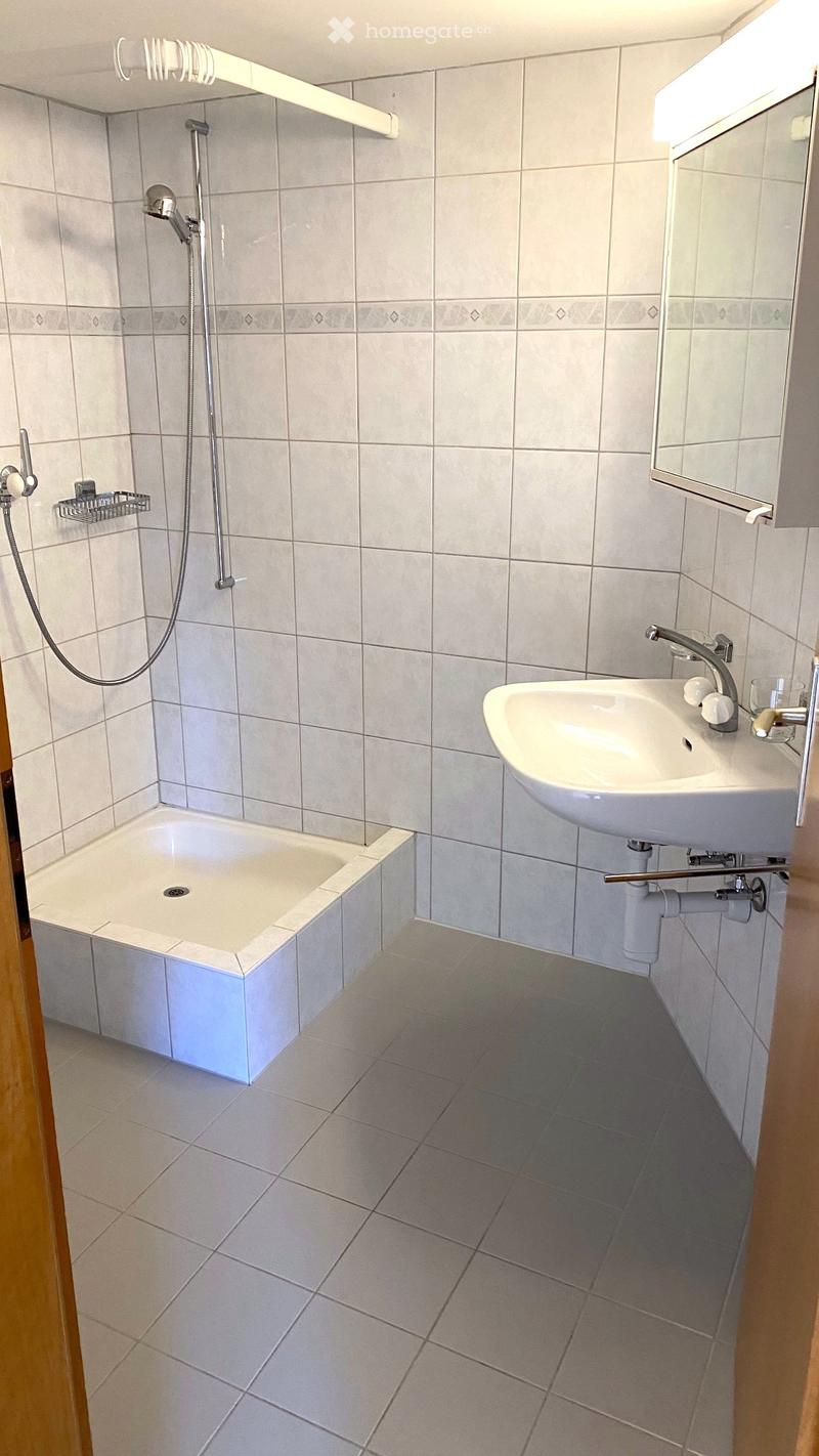 Modern bathroom with a bathtub, sink, and shower head. The walls and floor are tiled in light gray, and there is a mirror above the sink.