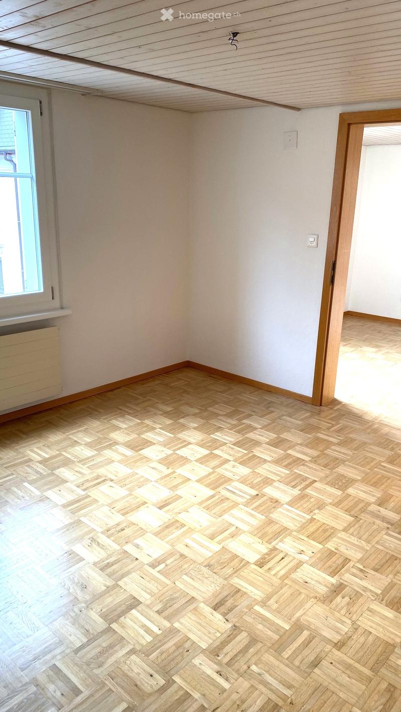 Empty room with wooden herringbone flooring, white walls, and a window with a radiator below it. The ceiling has exposed wooden beams.