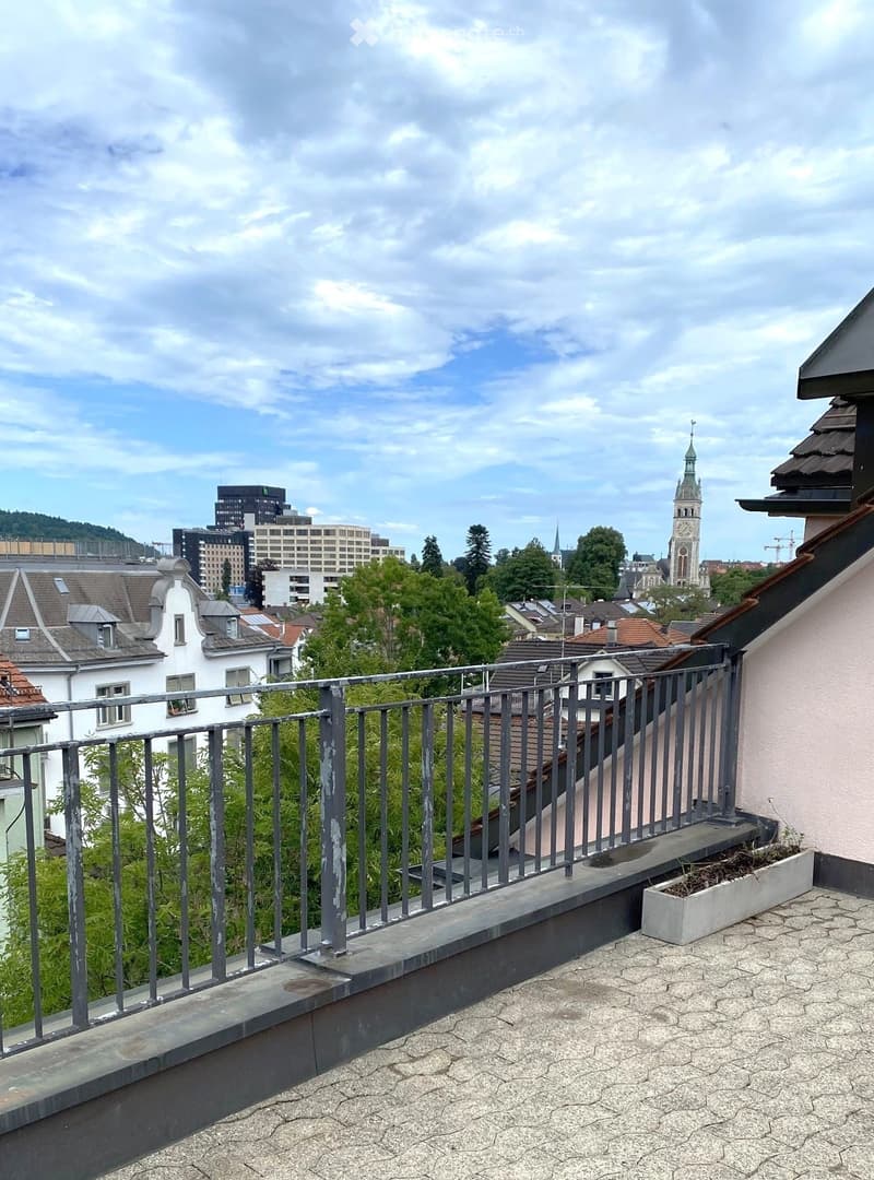 Balcony with metal railing, tiled roof surface, and a small planter, overlooking a cityscape with buildings and a church tower.