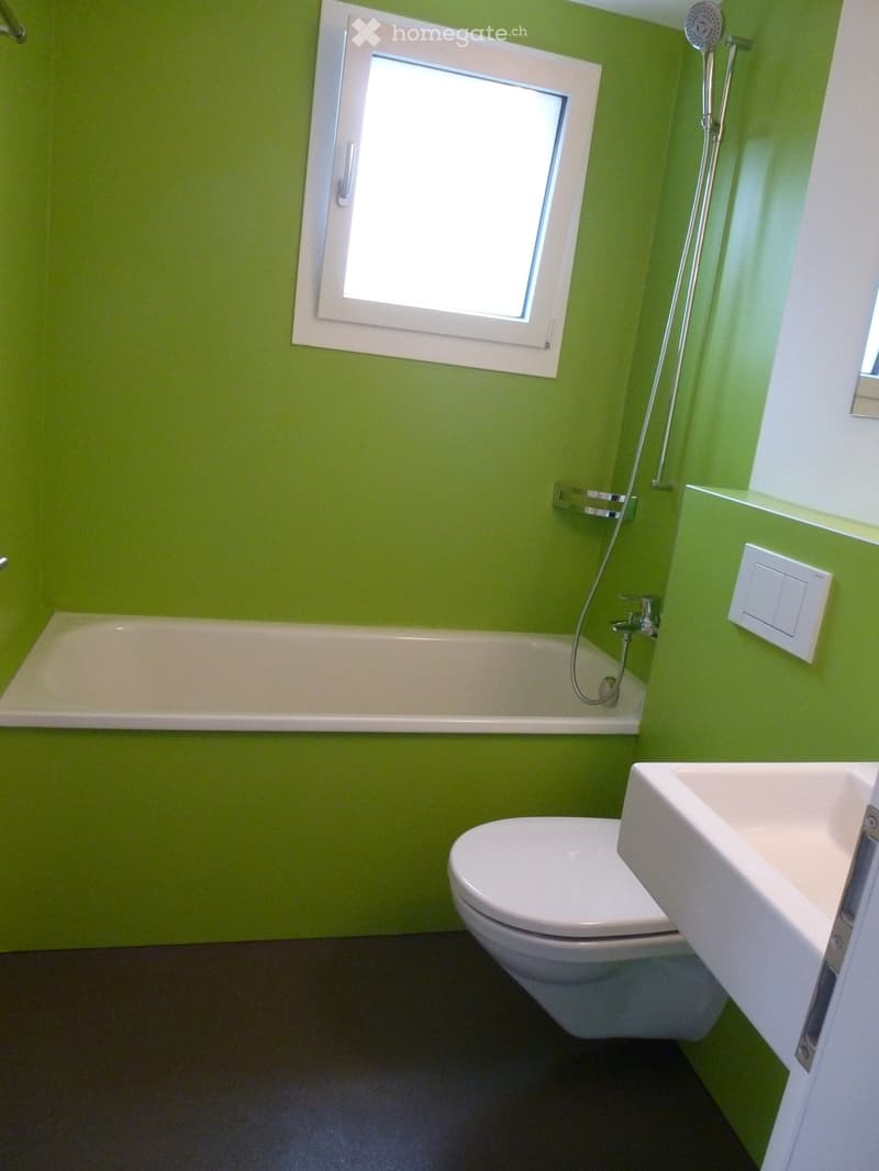 Modern bathroom with green walls, white fixtures including a bathtub, toilet, sink, and showerhead.