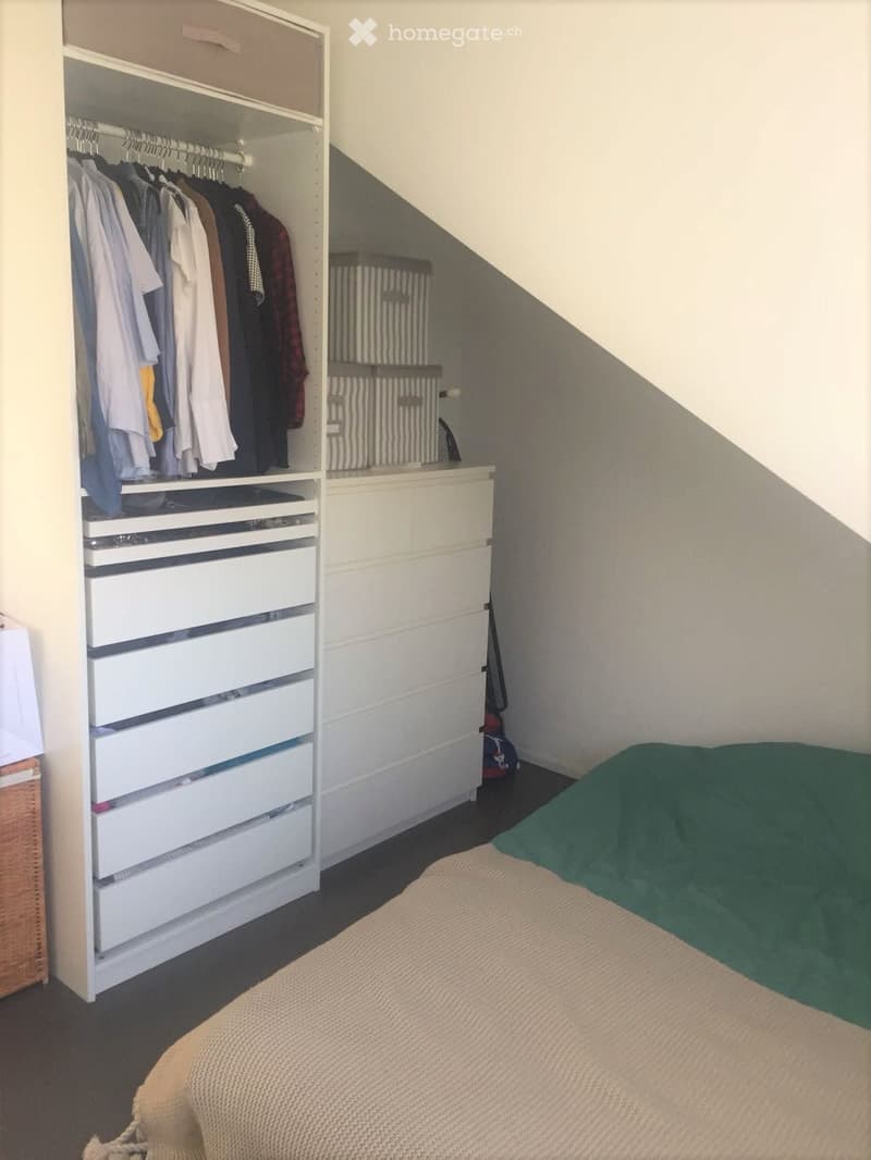 Bedroom with a neatly made bed, a large white wardrobe with hanging clothes and multiple drawers, and a slanted ceiling typical of an attic space.