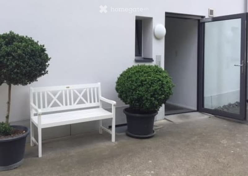 A small outdoor seating area with a white wooden bench, two potted shrubs, and a modern glass door leading inside.