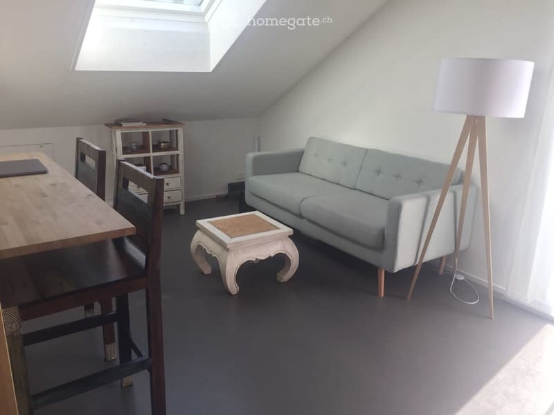 Bright attic living space with a grey sofa, wooden table, chairs, a small white shelf, and a floor lamp near a skylight window.