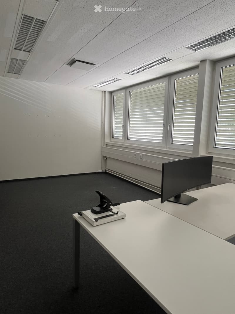 Office space with white walls, black carpet, large windows with blinds, a white desk with a stapler, and a monitor on a stand.