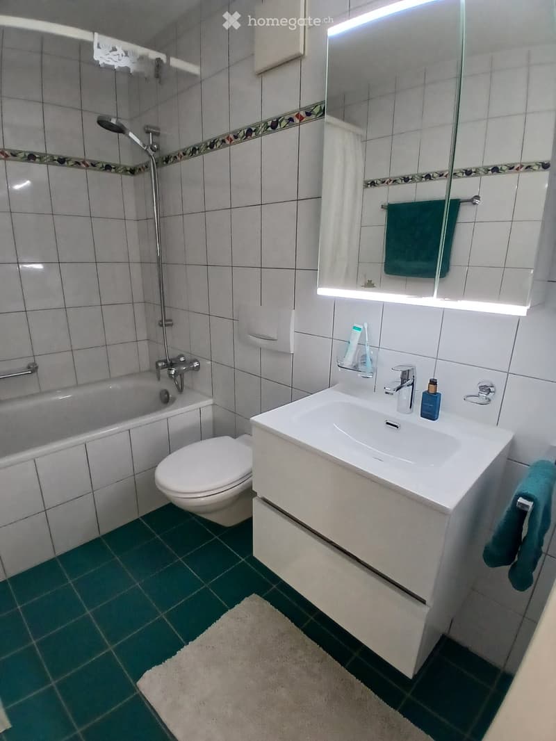 Modern bathroom with white tiles, a bathtub, toilet, sink, mirror, and a green towel. The floor is green tiled with a white rug in front of the sink.
