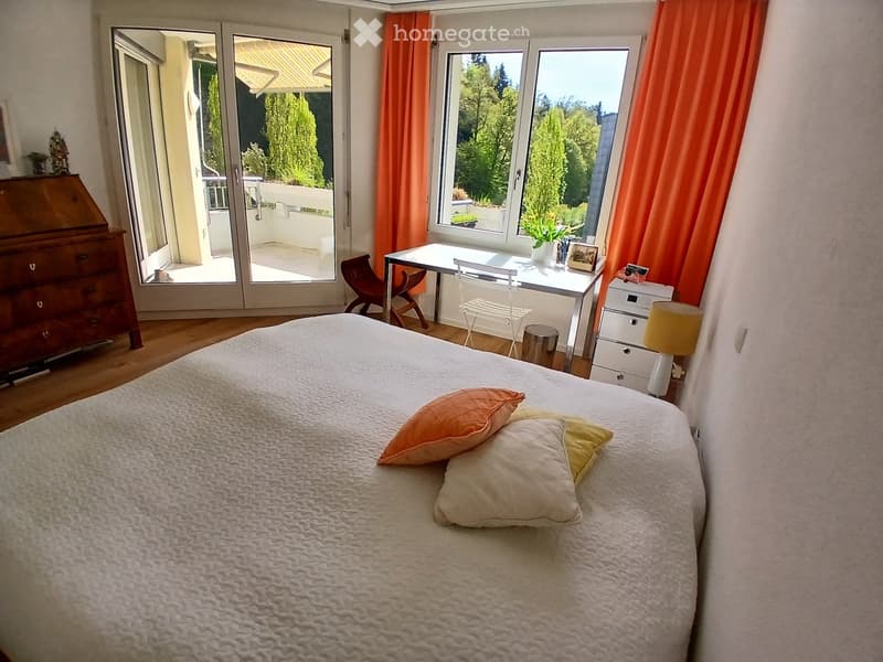 Bright bedroom with a large bed, pillows, a wooden chair, a small table, a lamp, and a cabinet. Large windows and a sliding glass door lead to a balcony.