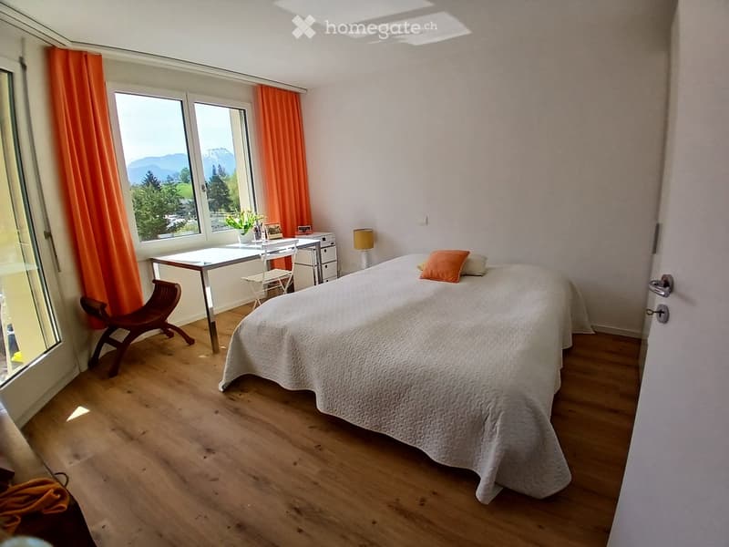 Bright bedroom with wooden flooring, large windows offering a scenic mountain view, a neatly made bed, a wooden chair, a small table with a lamp, and a potted plant on the windowsill.