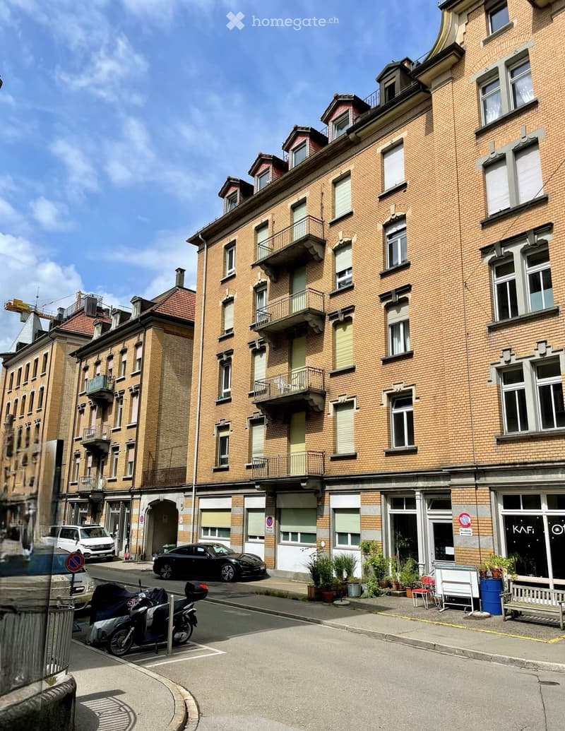 Multi-story brick building with multiple windows and balconies, located on a street with parked cars and a few outdoor plants.