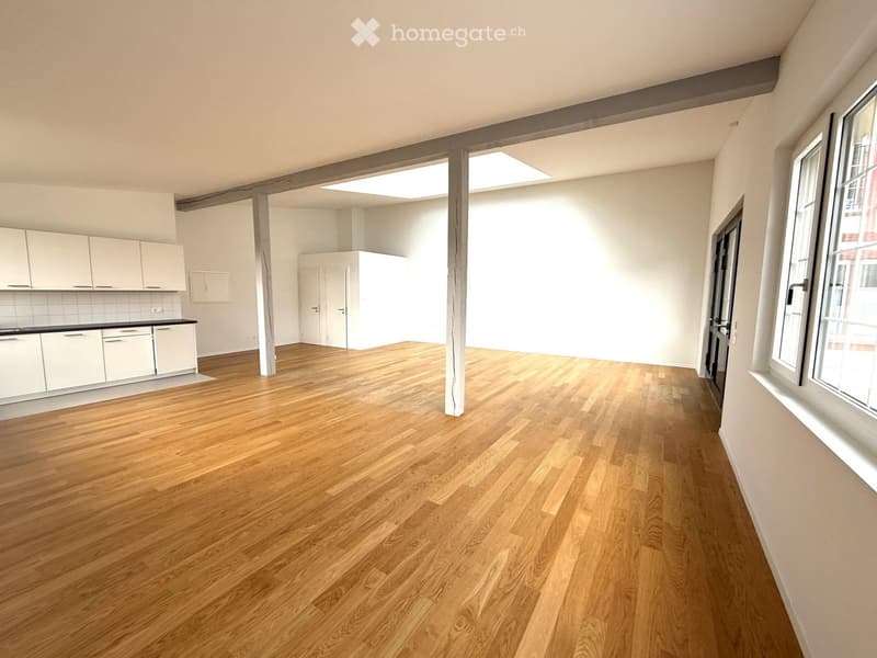 Empty room with hardwood floors, white walls, kitchen area with cabinets, and windows.