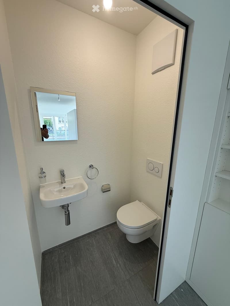 Modern, compact bathroom with a white sink, toilet, and mirror. Features dark tiled flooring and white walls.
