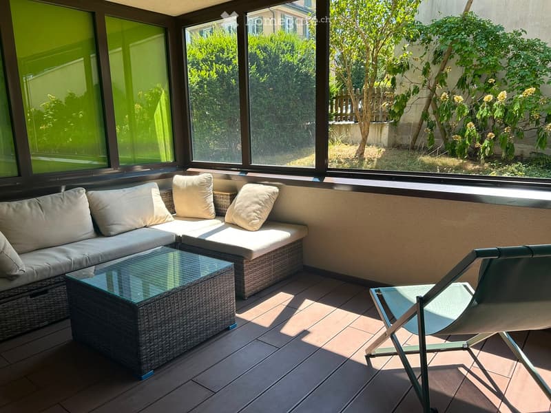 Balcony with a wicker sofa, glass coffee table, and a single chair, overlooking a garden with trees and shrubs.