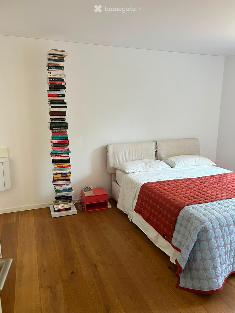 A tidy bedroom with a wooden floor, a neatly made bed with a red and blue quilt, a nightstand, and a tall stack of books against the wall.