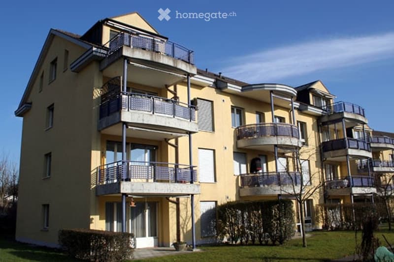 Modern multi-story apartment building with yellow facade, multiple balconies, and a well-maintained lawn in front.