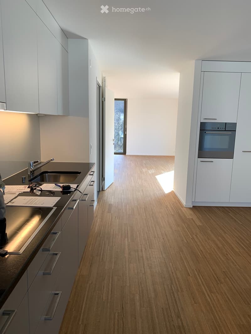 Modern kitchen with white cabinets, wooden flooring, stainless steel sink, and built-in oven. Countertop has a few items including a measuring tape and papers.