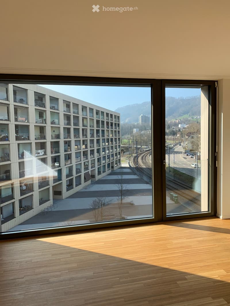 Empty room with wooden flooring and large windows offering a view of a modern building and surrounding landscape.
