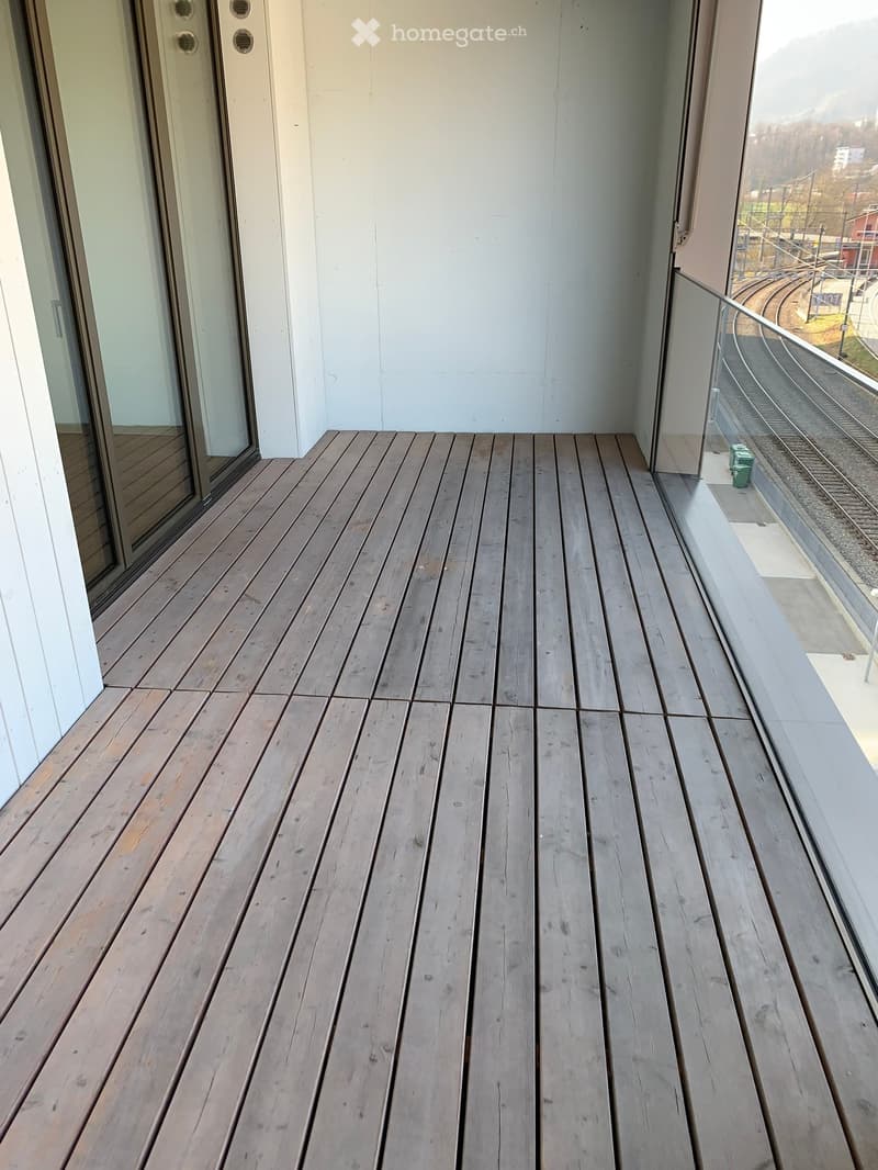 Wooden deck balcony with glass railing, overlooking a train track and outdoor area.