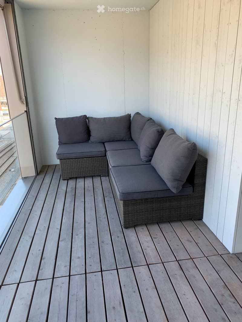 Wooden balcony with a grey wicker sofa and matching cushions, white walls, and a glass railing.