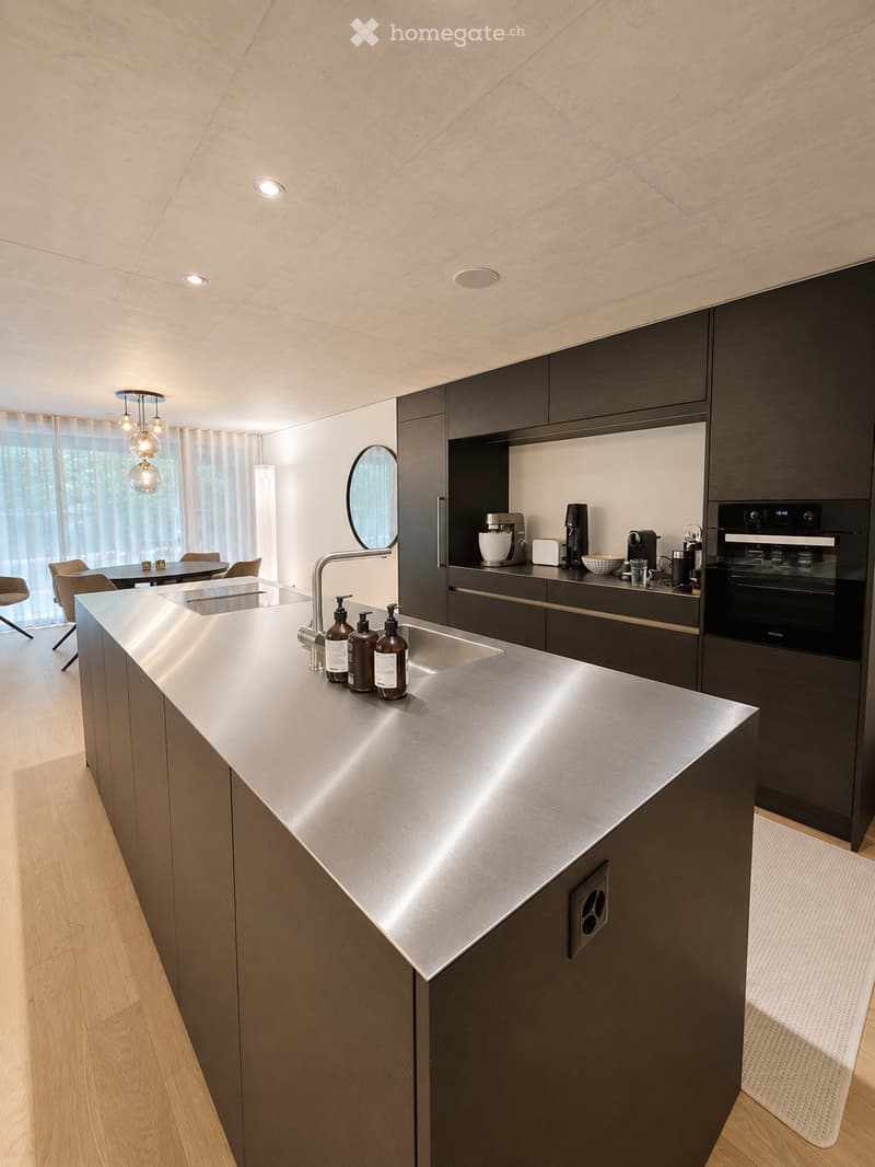 Modern kitchen with stainless steel island, dark wood cabinets, and various appliances including a coffee maker, blender, and oven. The space features a sleek design with pendant lights and a large window with sheer curtains.
