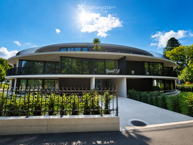 Modern, curved two-story building with large glass windows and balconies, surrounded by greenery and a metal fence.