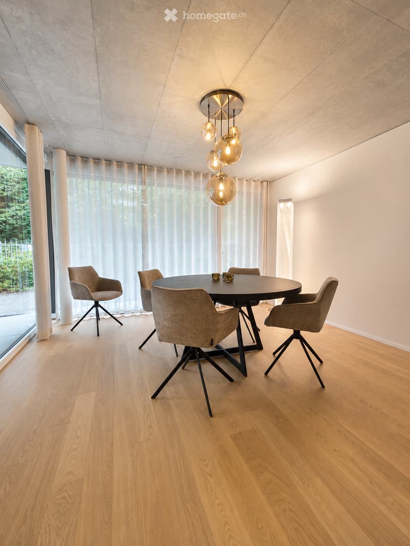 Modern dining room with a round black dining table, four grey upholstered chairs, pendant light fixture, large window with sheer curtains, and wooden flooring.