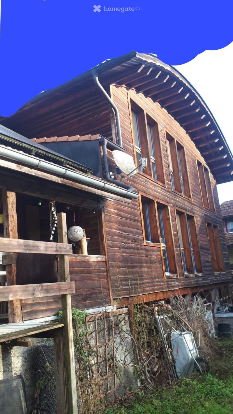 A two-story wooden house with a curved roof, multiple windows, and a balcony on the upper level. The house has a stone foundation and is surrounded by overgrown vegetation.
