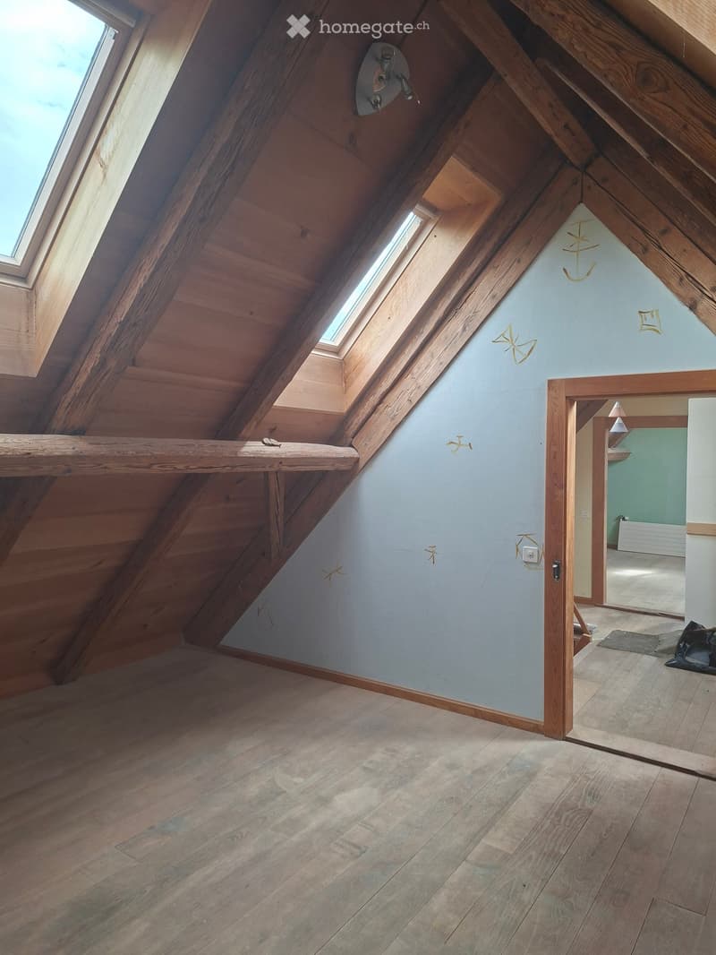 An attic space with wooden beams, skylights, and a built-in wooden shelf. The floor is made of light-colored wood planks, and the walls are painted a soft blue. A doorway leads to another room with a visible green wall and a white cabinet.