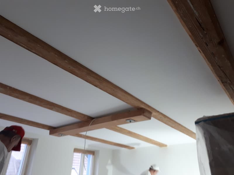 Empty room with exposed wooden beams on the ceiling, white walls, and windows allowing natural light to enter.
