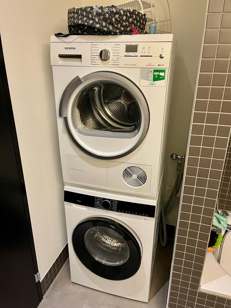 A compact bathroom with a stacked Siemens washing machine and dryer unit, featuring a control panel and a small shelf above the dryer holding a bag and other items.