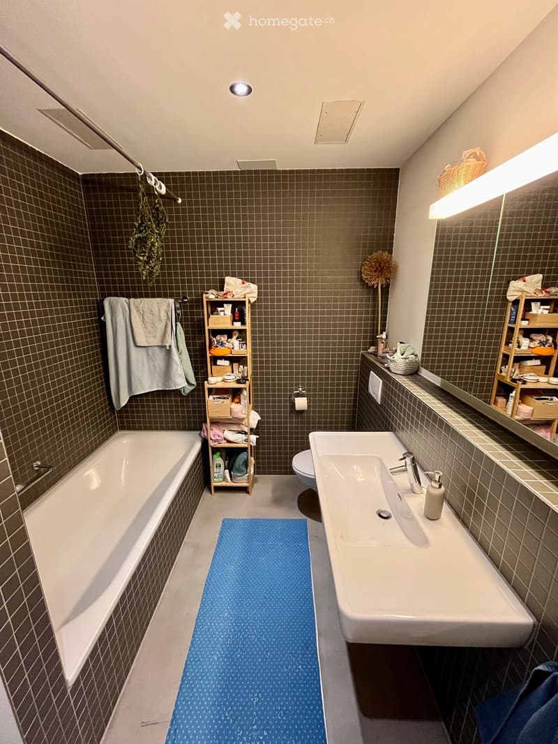 Bathroom with a bathtub, sink, toilet, and shelves holding toiletries. Features a blue bath mat on the floor and towels hanging on the wall.