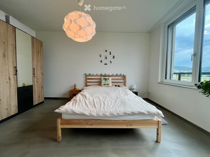 Modern bedroom with a wooden bed, white bedding, nightstand, lamp, and a large window offering a scenic view.