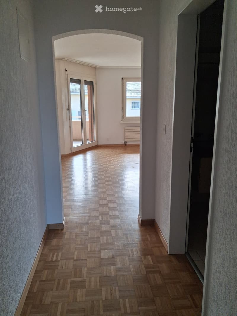 Empty hallway with wooden herringbone flooring, light-colored walls, and an arched opening leading to another room with a sliding glass door and visible outdoor area.