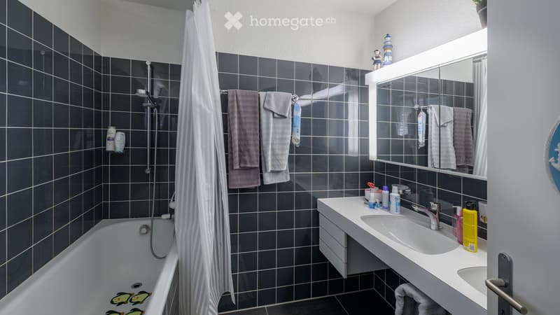Modern bathroom with dark tiled walls, a bathtub, a sink with a mirror, and towels hanging on a rack. The shower area has a curtain and a showerhead.