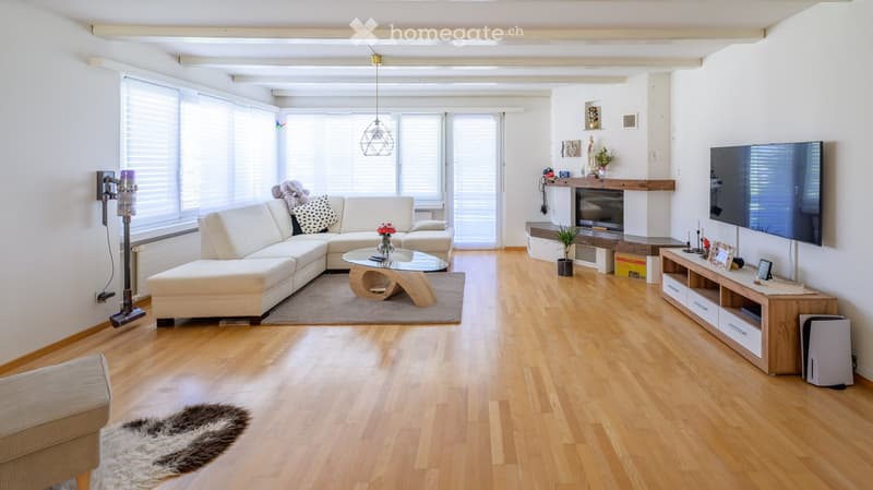 Modern living room with light wooden flooring, large windows, a white sectional sofa, a glass coffee table, a fireplace, a TV stand, and decorative items.