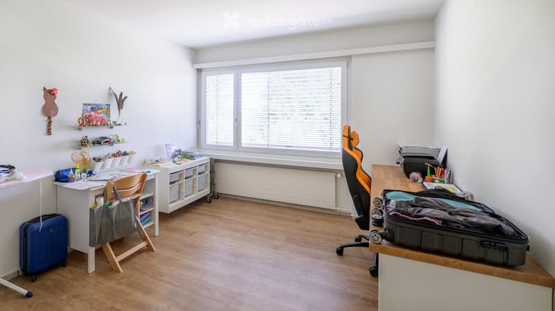 A well-lit room with wooden flooring, large window with blinds, and various furniture including a desk, shelves, and a gaming chair.