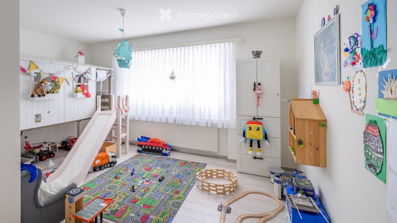 Brightly lit children's room with a play slide, toy cars, train set, and various toys. Features a sofa, table, and colorful artwork on the walls.