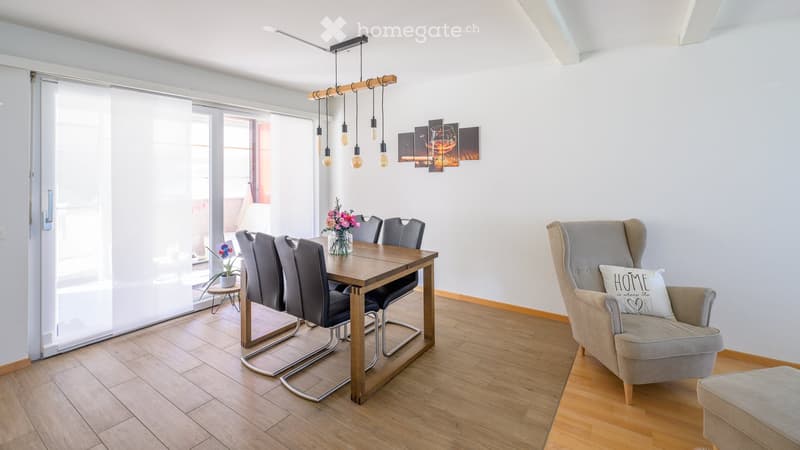 Bright dining area with wooden table, six chairs, vase with flowers, grey armchair, and artwork on wall.