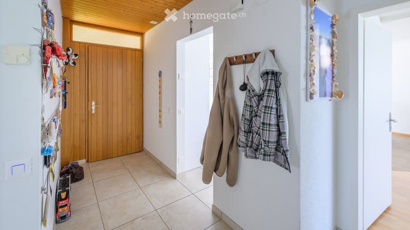 A well-lit hallway with tiled flooring, white walls, and a wooden ceiling. A coat rack with jackets, a small shelf with decorative items, and a toy car on the floor are visible.
