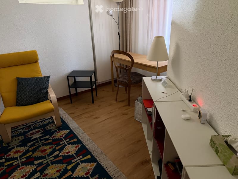 A cozy, well-lit room with a yellow armchair, a small side table, a desk with a chair, a lamp, and a shelf with various items.