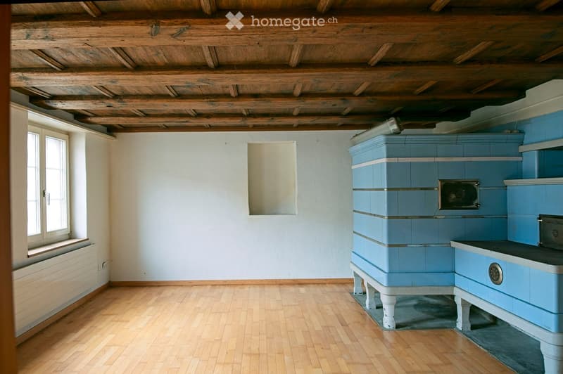 Empty room with wooden floor, exposed wooden ceiling beams, light blue tiled stove, and a window with white frame.