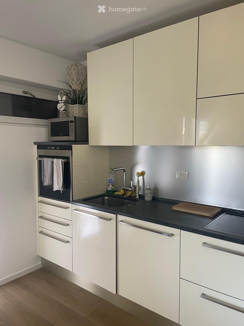 Modern kitchen with white cabinets, stainless steel appliances, and a black countertop. Features an oven, microwave, sink, and various small items on the counter.